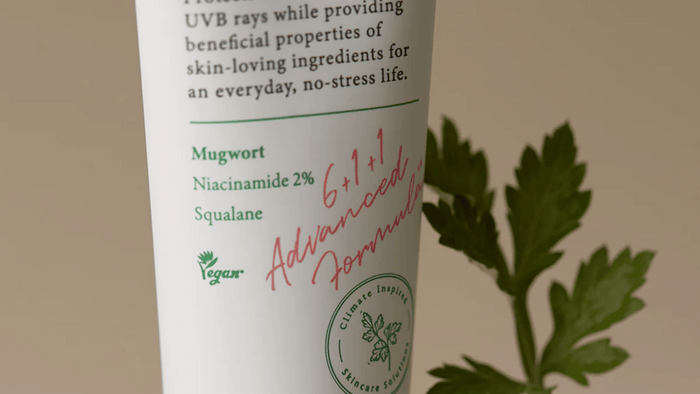 En närbild av en vegansk hudvårdsprodukt med text som lyfter fram ingredienser som mugwort, niacinamid 2 % och squalane. En grön växt visas i bakgrunden, vilket förstärker produktens naturliga och miljövänliga profil.