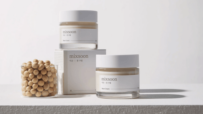 Två burkar av Mixsoon Bean Cream står på en ljus yta med en glasbehållare fylld med beige bönor och en produktkartong bredvid. Enkel och ren design med tydliga etiketter. Bakgrunden är neutral för att fokusera på produkterna.