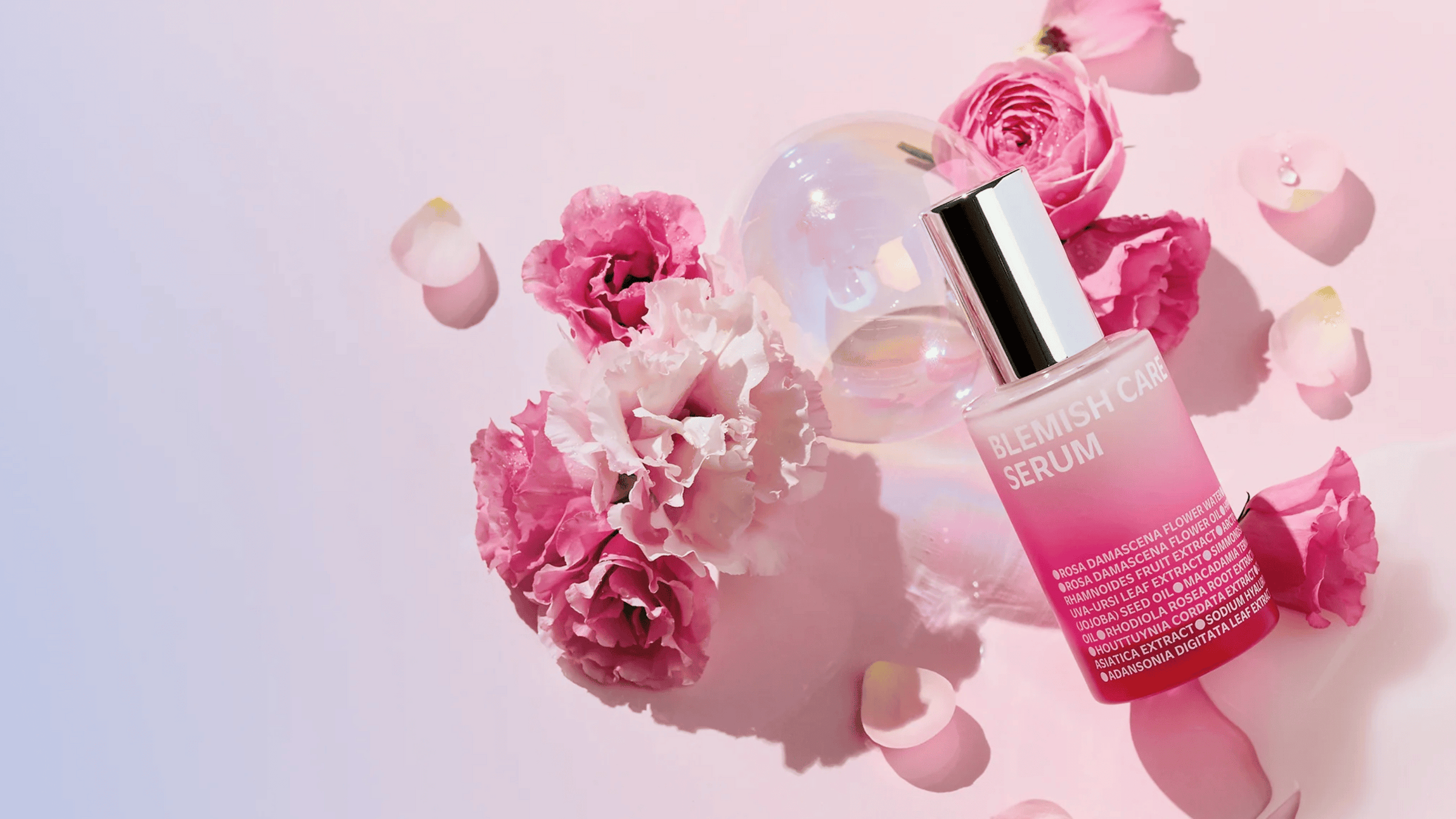 En flaska Blemish Care Serum ligger på en rosa bakgrund omgiven av rosa och vita blommor samt rosenblad. Flaskan har en silverfärgad kork och ett övertonat rosa innehåll, med produktinformation tryckt i vitt på framsidan.