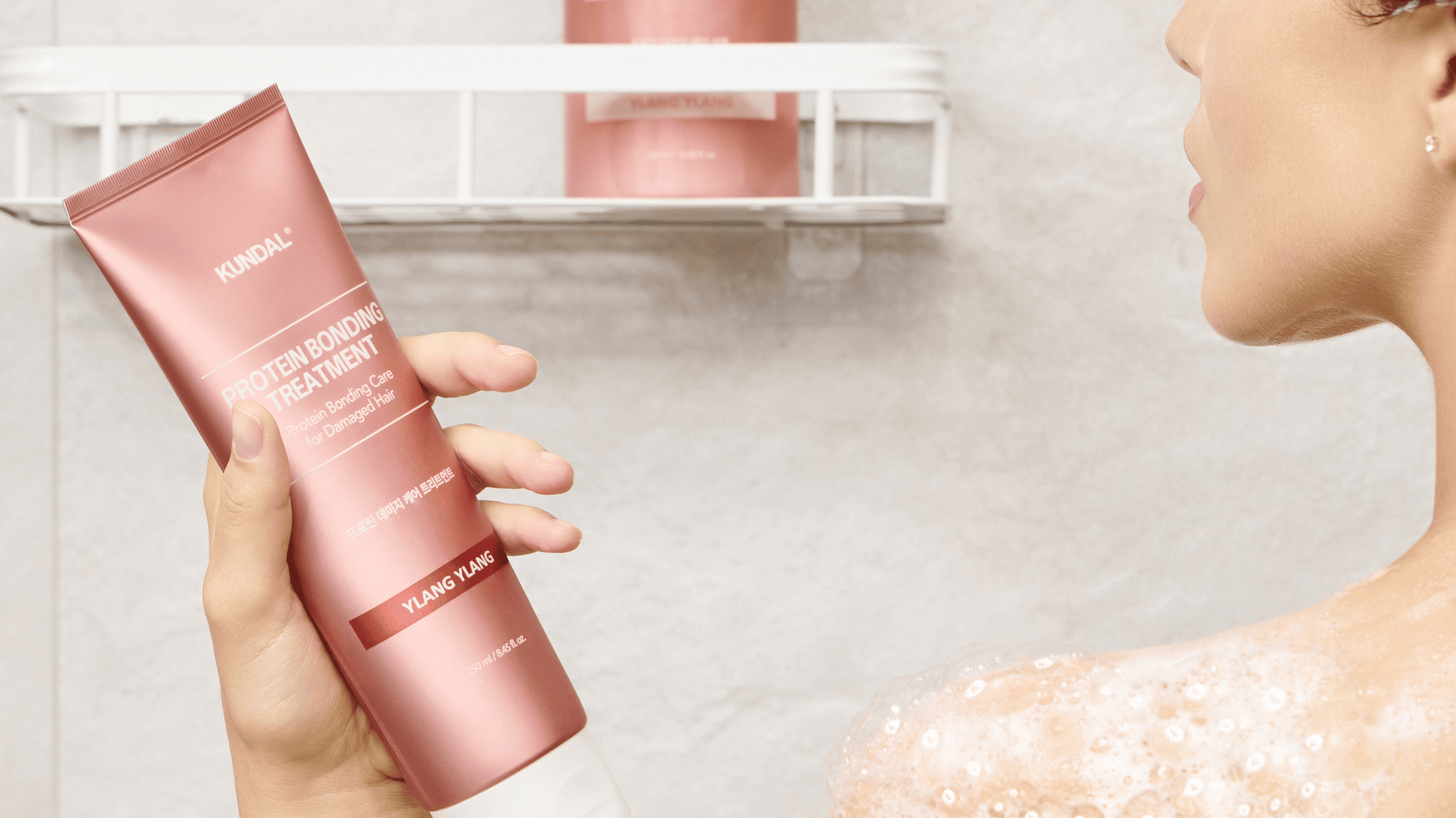 Person i duschen håller KUNDAL Protein Bonding Treatment Ylang Ylang, en hårbehandling för skadat hår, med lödder på huden och matchande flaska i hyllan.