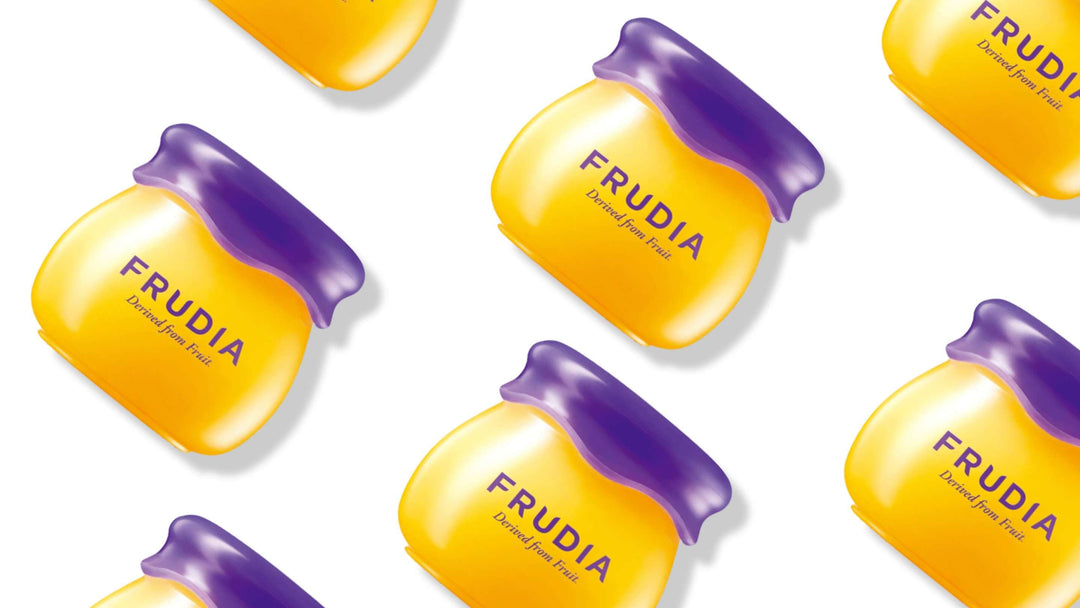 Bilden visar FRUDIA's hudvårdsprodukter i form av små, färgglada behållare som liknar frukt, placerade naturligt bland gröna blad och blåbär. Designen antyder naturliga ingredienser och produkternas fräschhet.