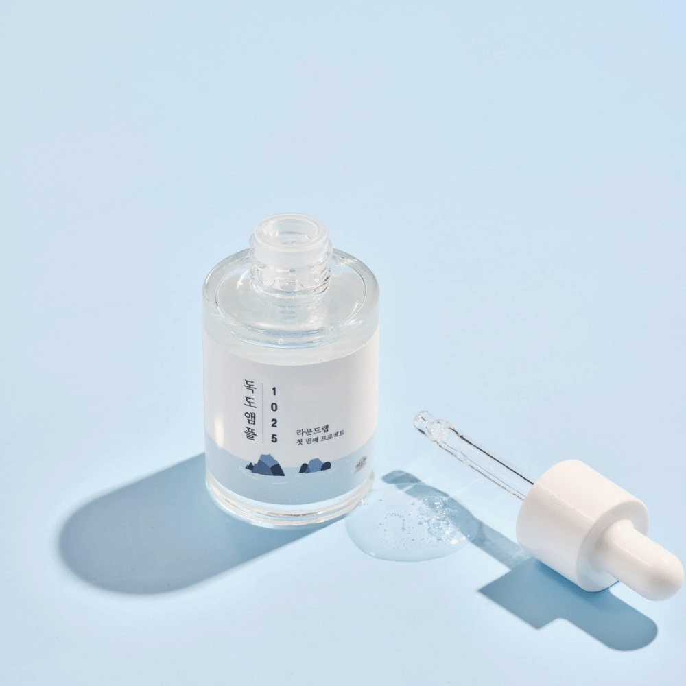 Une bouteille ouverte de 1025 Dokdo Ampoule avec une pipette à côté, sur un fond bleu créant une ombre douce.