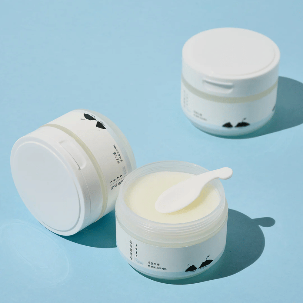 Trois pots de 1025 Dokdo Cleansing Balm, un ouvert avec une spatule dessus, sur un fond bleu clair avec des ombres nettes.