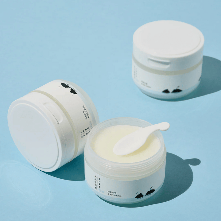 Trois pots de 1025 Dokdo Cleansing Balm, un ouvert avec une spatule dessus, sur un fond bleu clair avec des ombres nettes.