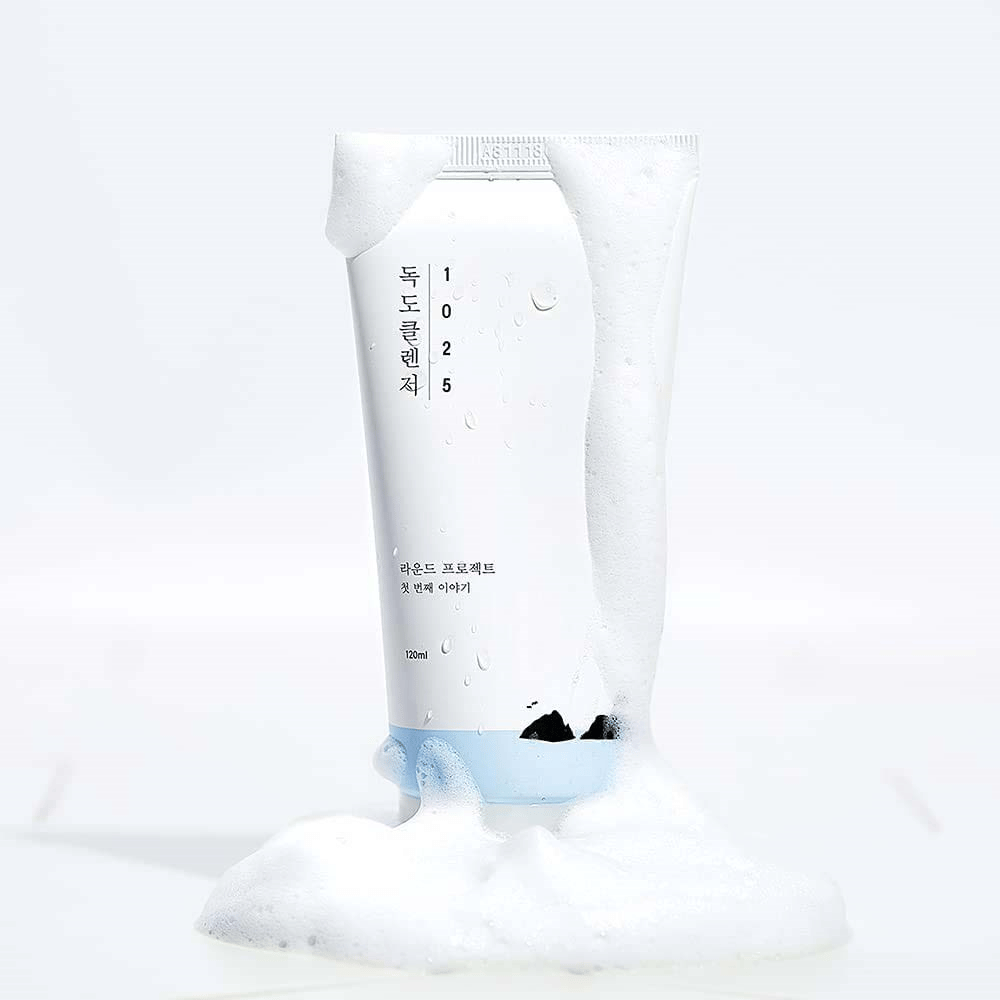 1025 Dokdo Cleanser dans un tube blanc couvert de mousse, avec des perles d'eau et de la mousse autour du fond, isolé sur fond clair.