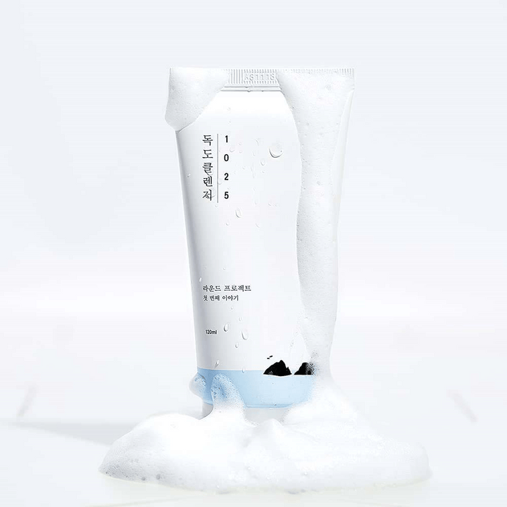 1025 Dokdo Cleanser dans un tube blanc couvert de mousse, avec des perles d'eau et de la mousse autour du fond, isolé sur fond clair.