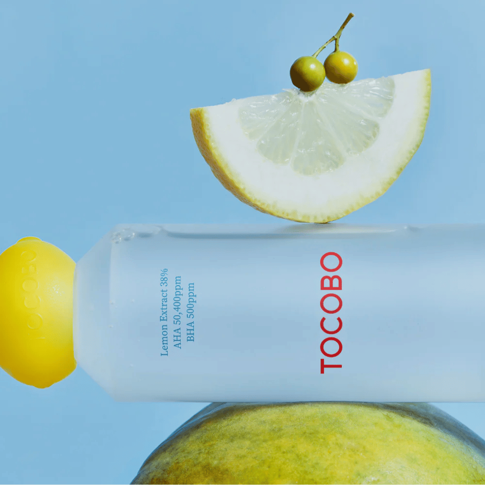 Bouteille avec TOCOBO AHA BHA Lemon Toner, décorée d'un bouchon en forme de citron. Une tranche de citron et de petites baies vertes équilibrent au-dessus de la bouteille sur un fond bleu.