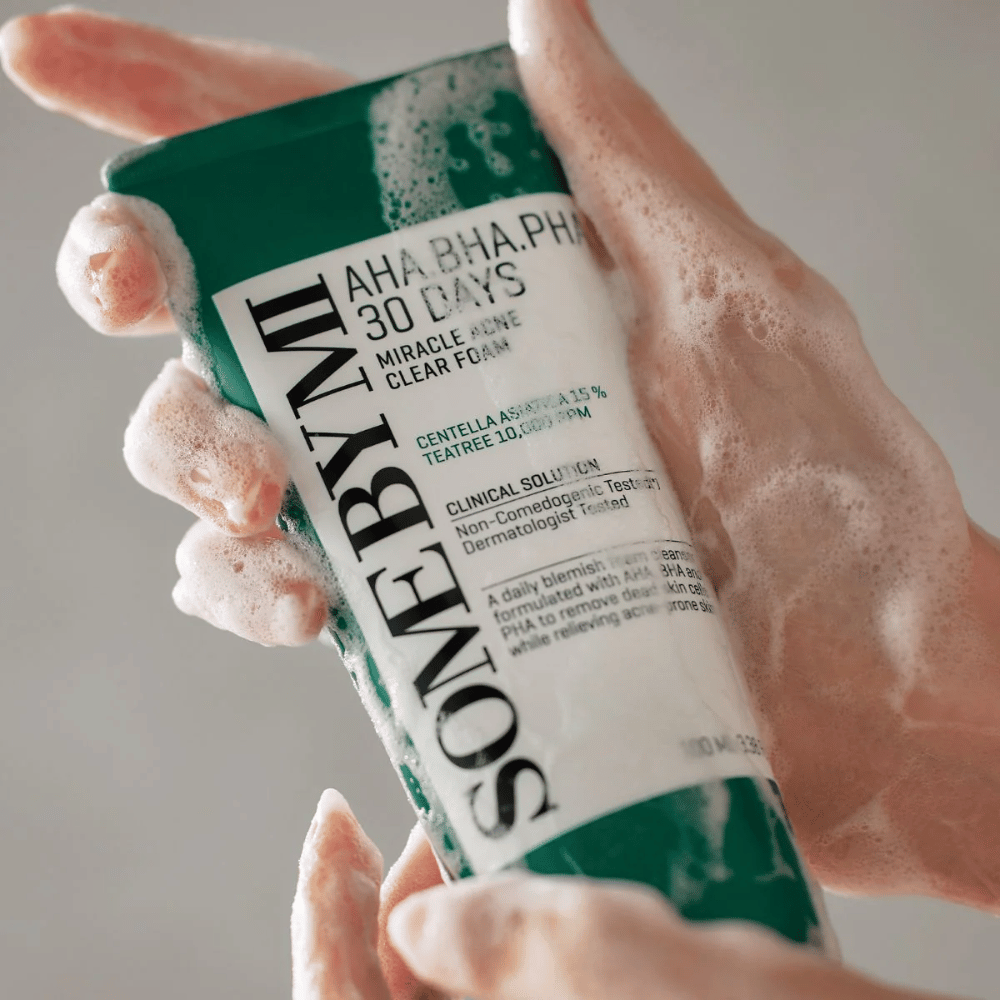 Une personne tient un tube de "SOME BY MI AHA, BHA, PHA 30 Days Miracle Acne Clear Foam" avec un contenu moussant sur les mains. Le produit est destiné à un usage quotidien pour combattre l'acné et purifier les pores grâce à ses ingrédients actifs comme la centella, l'huile de tea tree et des acides tels que AHA, BHA et PHA.