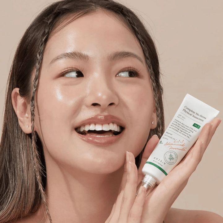 L'image montre une jeune femme souriante tenant un tube de AXIS-Y Complete No-Stress Physical Sunscreen. Elle a un aspect radieux et naturel avec un maquillage minimal, ce qui met en valeur le produit qu'elle présente.