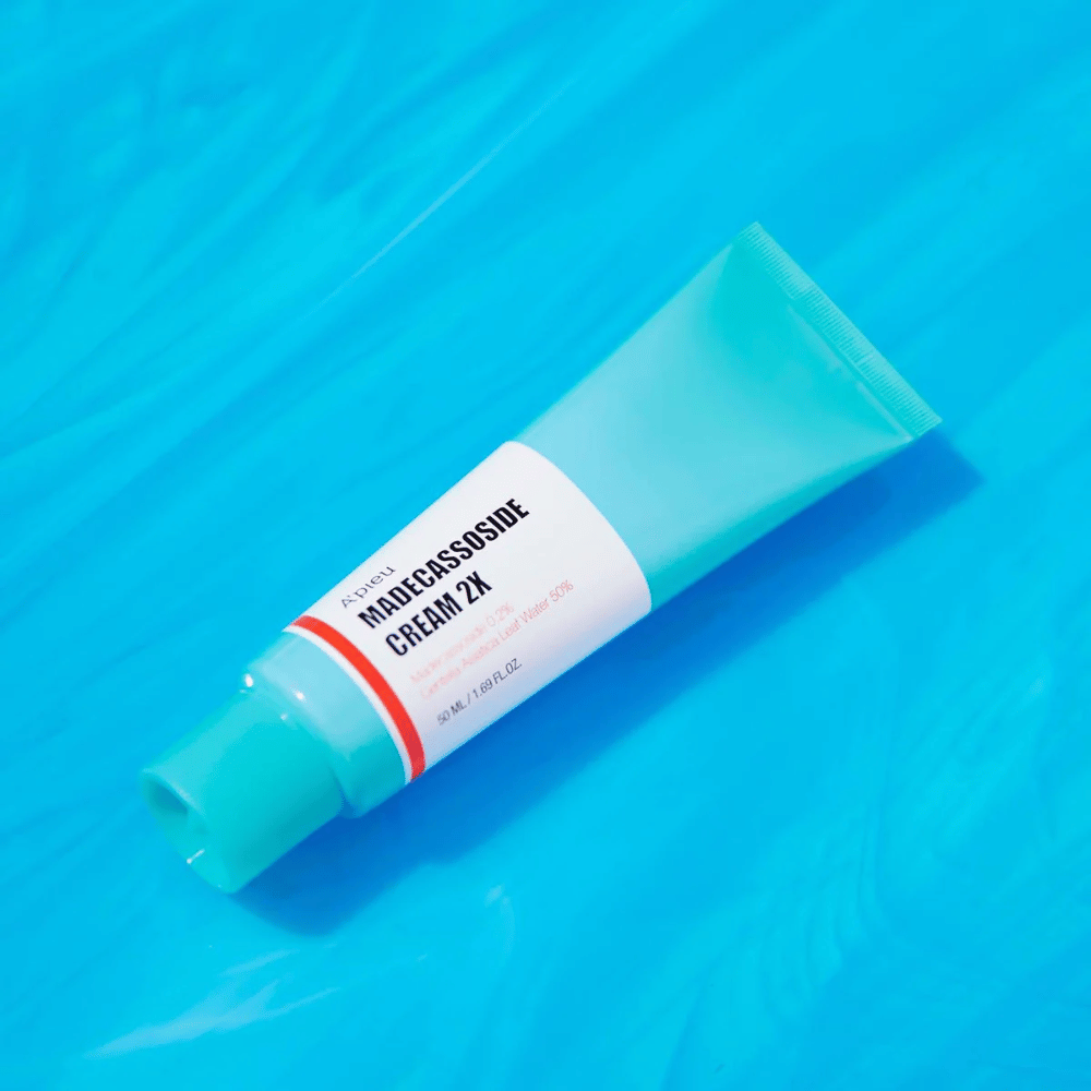 Un tube A'PIEU Madecassoside Cream 2X est posé en diagonale sur un fond bleu avec des plis semblables à de l'eau. Le tube a un corps blanc avec un bouchon turquoise clair et une étiquette rouge à orange où le nom du produit et l'ingrédient actif Madecassoside 2% sont clairement inscrits. L'étiquette indique également la quantité qui est de 50 ml / 1.8 fl oz. La présentation lumineuse et colorée donne une sensation fraîche et énergique.
