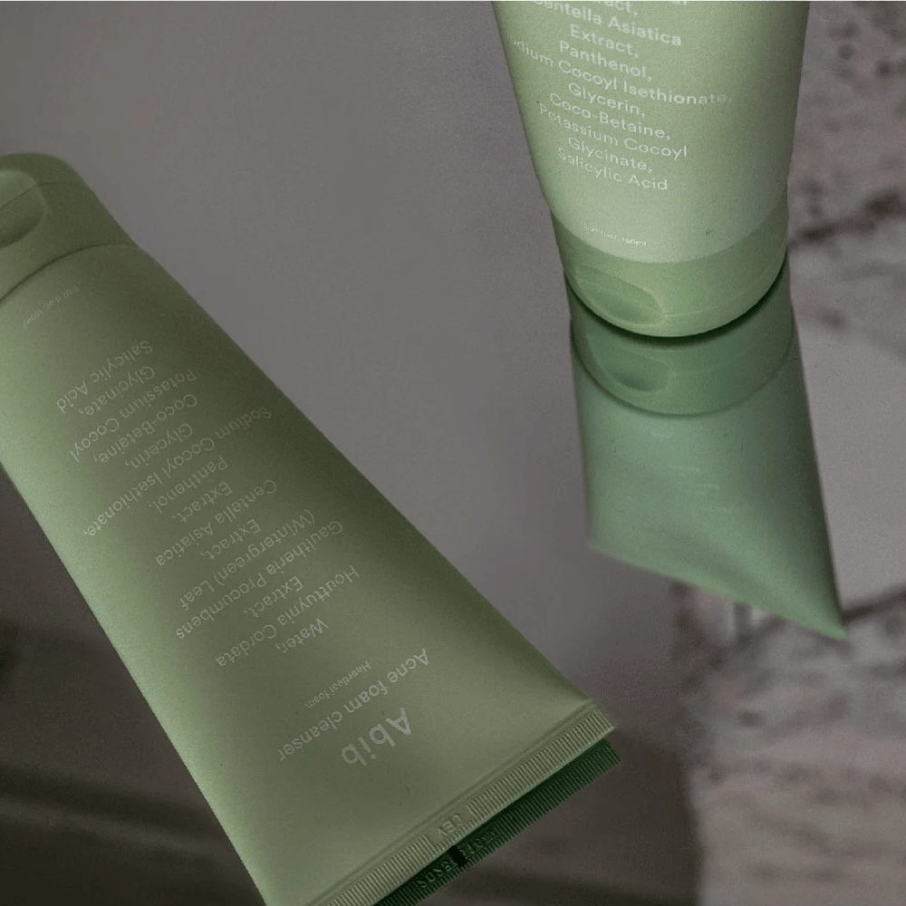 Tube vert de Abib Acne Foam Cleanser Heartleaf allongé sur une table miroir avec le devant du produit et le texte reflétés. Des ingrédients comme l'acide salicylique, la centella et l'extrait de feuille de cœur sont clairement visibles sur l'emballage.