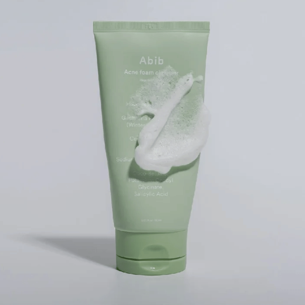 Tube vert avec Abib Acne Foam Cleanser Heartleaf, recouvert d'une mousse blanche illustrant l'effet nettoyant moussant. Le produit contient de l'extrait de feuille de cœur et de l'acide salicylique pour les peaux sujettes à l'acné.