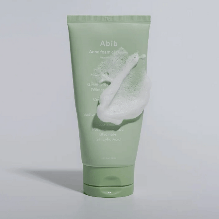 Tube vert avec Abib Acne Foam Cleanser Heartleaf, recouvert d'une mousse blanche illustrant l'effet nettoyant moussant. Le produit contient de l'extrait de feuille de cœur et de l'acide salicylique pour les peaux sujettes à l'acné.