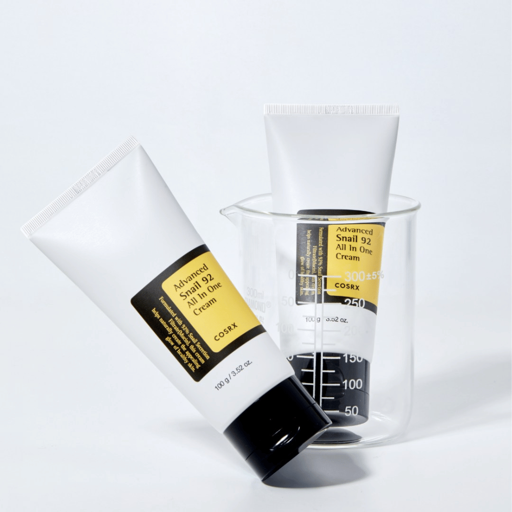 Un tube de COSRX Advanced Snail 92 All in One Cream allongé à côté d'un cylindre de mesure transparent renversé sur un fond blanc.