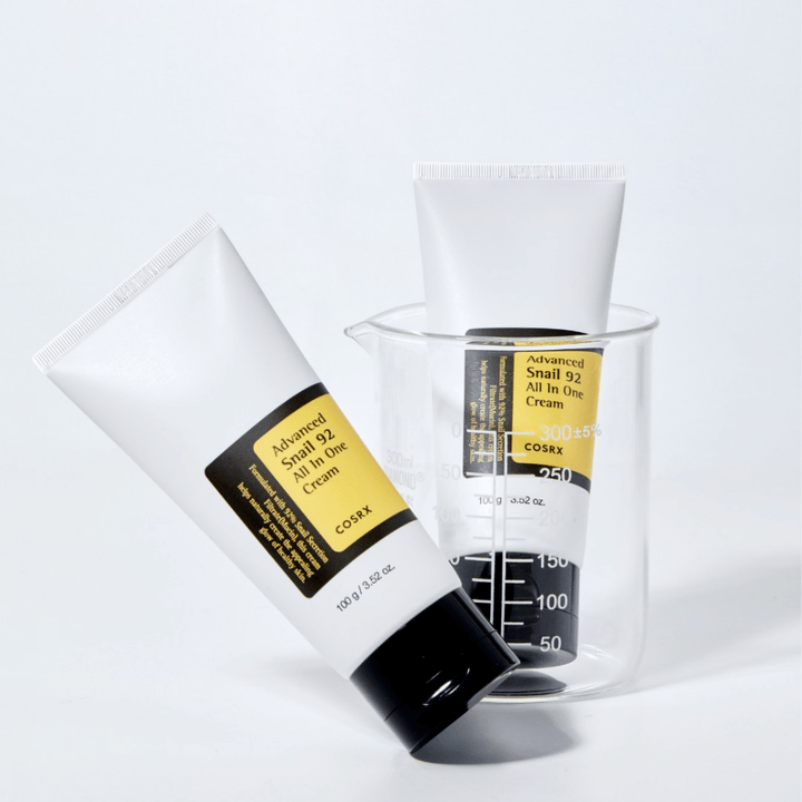 Un tube de COSRX Advanced Snail 92 All in One Cream allongé à côté d'un cylindre de mesure transparent renversé sur un fond blanc.