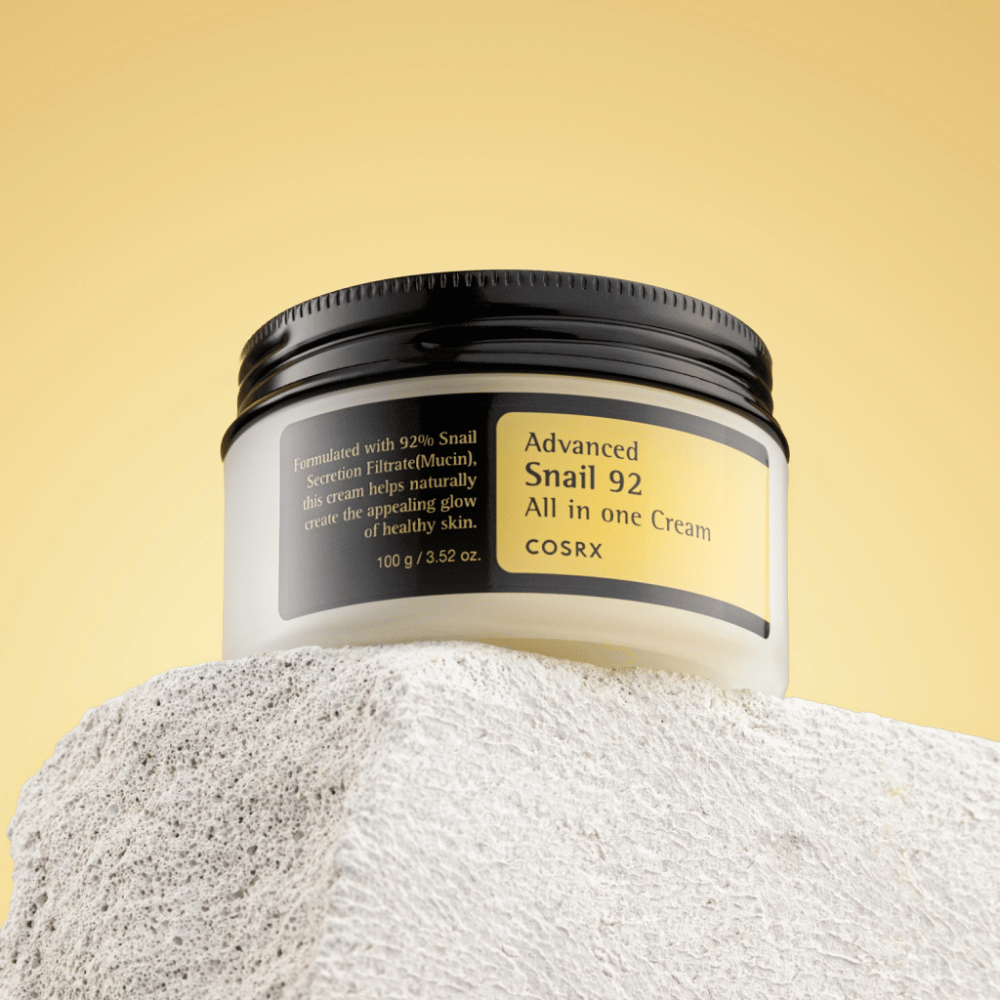 Un pot de COSRX Advanced Snail 92 All in one Cream placé sur une surface rocheuse avec un fond jaune.