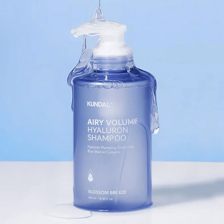 KUNDAL Airy Volume Hyaluron Shampoo Blossom Breeze dans un flacon pompe bleu, avec de l'acide hyaluronique et Blue Marine Complex, photographié avec de l'eau courante.