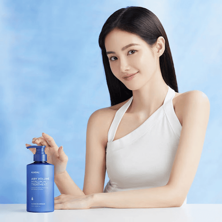 Femme avec de longs cheveux brillants pose avec KUNDAL Airy Volume Hyaluron Treatment Blossom Breeze dans un flacon pompe bleu.