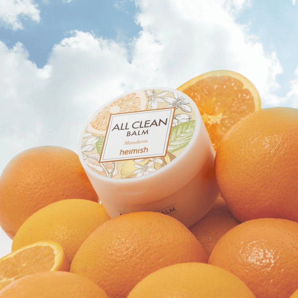 Pot de Heimish All Clean Balm Mandarin placé sur un tas d'oranges, avec un ciel bleu en arrière-plan.