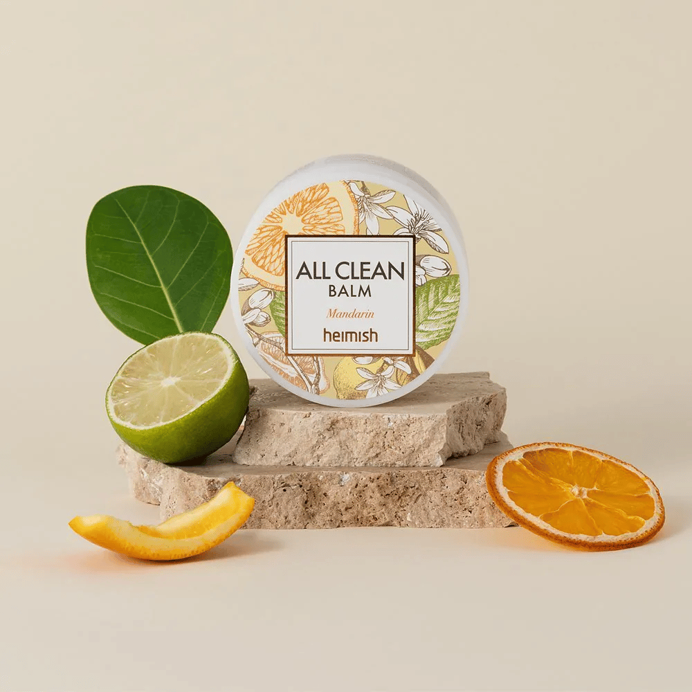 Pot de Heimish All Clean Balm Mandarin placé sur des pierres, entouré d'écorces de mandarine et de feuilles de citronnier, devant un fond clair.