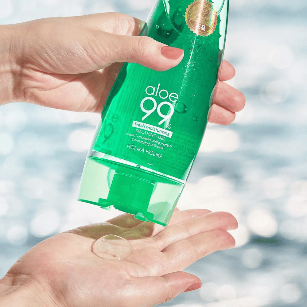 Une main tient un tube vert avec Holika Holika Aloe 99% Soothing Gel, tandis qu'une autre main attrape une noisette de gel qui est distribuée. L'arrière-plan est flou et montre un motif aquatique scintillant.