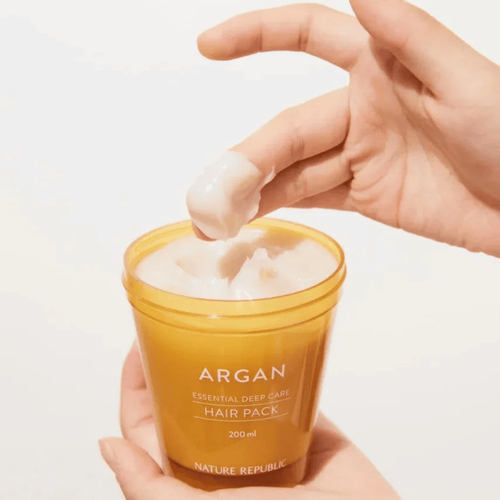 Une main prend la formule crémeuse de Nature Republic Argan Essential Moisture Repairing Hair Mask – nutrition intensive pour cheveux secs.