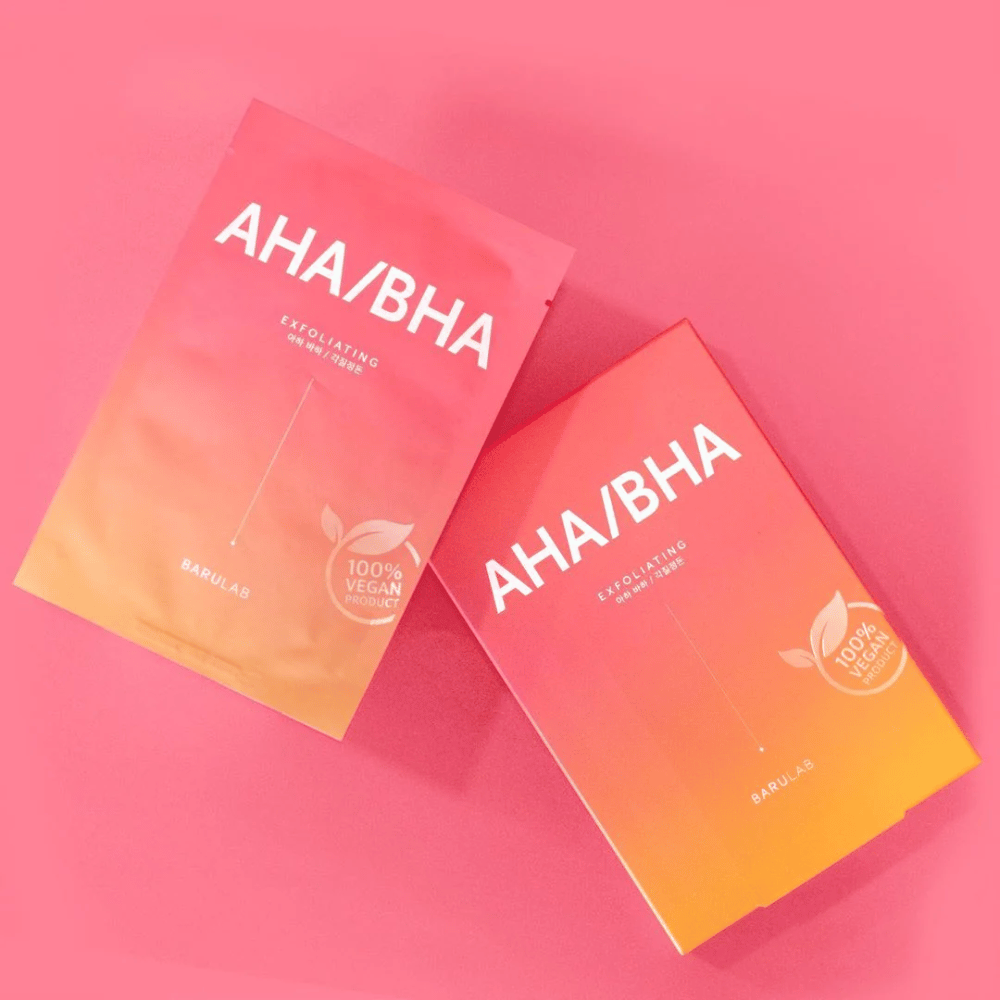 Deux produits de BARULAB, un dans un sac et un dans une boîte, tous deux marqués "AHA/BHA" et "Exfoliating" sur un fond rose. Les produits sont marqués comme "Produit 100% Végétalien".