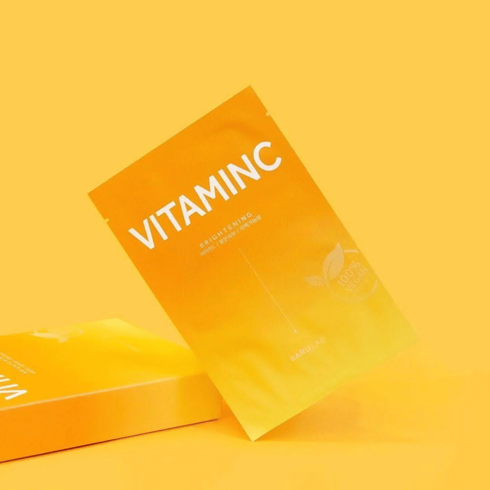Une pile de paquets de masques "VITAMINC Brightening" BARULAB, tous marqués comme "100% Vegan Product". Les emballages ont une teinte de couleur dans différentes nuances d'orange et de jaune, placés sur un fond jaune uni.