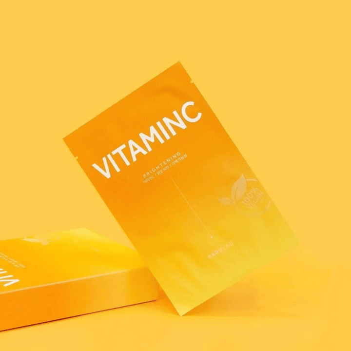 Une pile de paquets de masques "VITAMINC Brightening" BARULAB, tous marqués comme "100% Vegan Product". Les emballages ont une teinte de couleur dans différentes nuances d'orange et de jaune, placés sur un fond jaune uni.