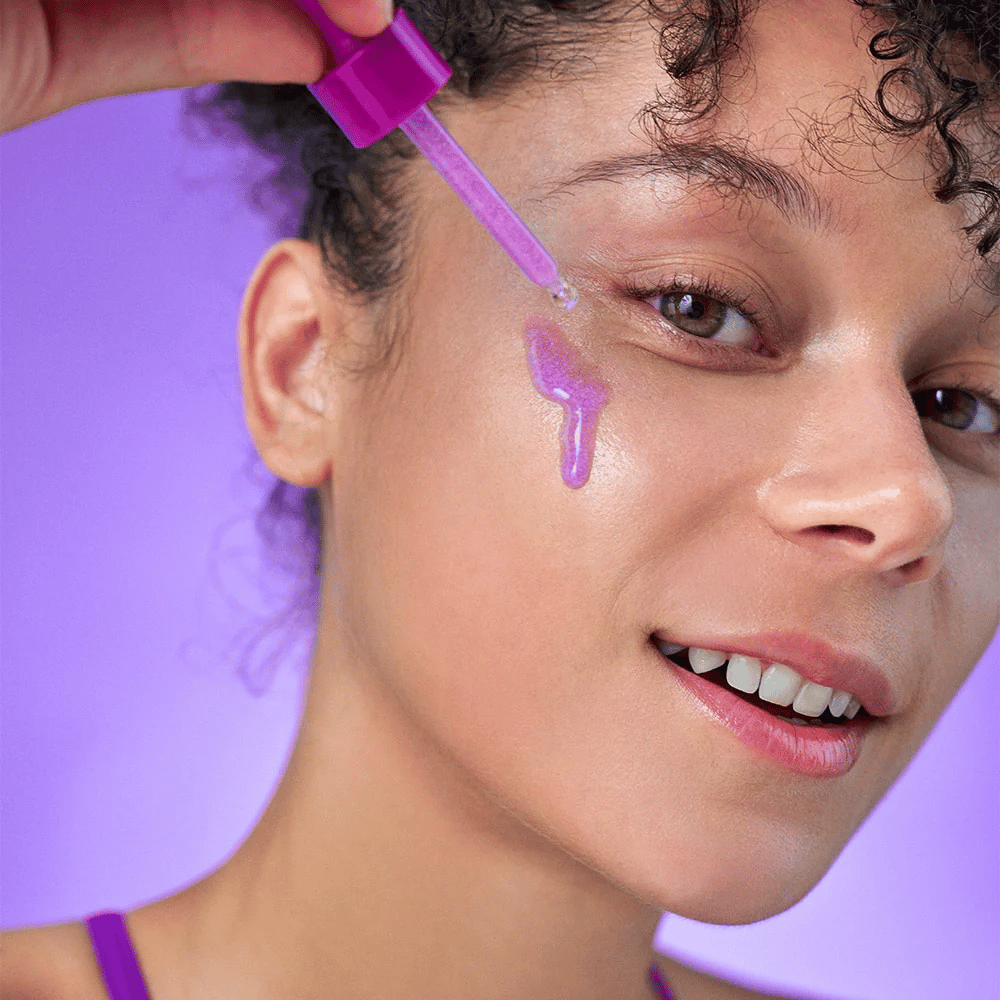 Femme souriante appliquant un sérum irisé violet sur la peau avec une pipette. Teint éclatant et fond violet assorti à la teinte du produit.
