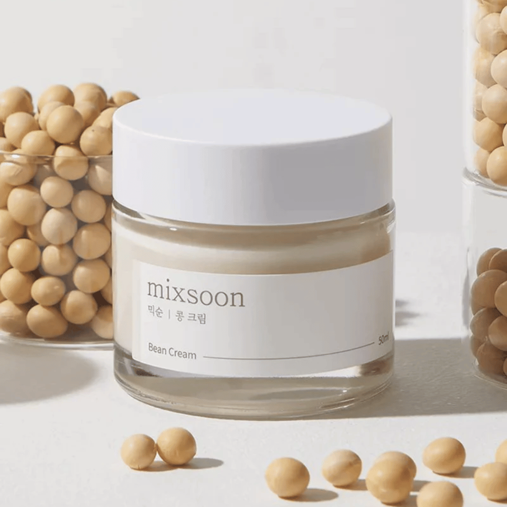 Un pot transparent avec une étiquette et un couvercle blancs, étiqueté "mixsoon 팥 크림" et "Bean Cream" avec un volume indiqué de "50ml". Le pot contient une crème jaune clair et est placé sur une surface claire. À côté du pot se trouve un récipient en verre rempli de haricots brun clair qui ont été renversés sur la surface, suggérant que la crème contient un extrait de haricot. L'arrière-plan et l'éclairage créent une atmosphère chaleureuse et naturelle.