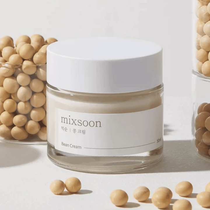 Un pot transparent avec une étiquette et un couvercle blancs, étiqueté "mixsoon 팥 크림" et "Bean Cream" avec un volume indiqué de "50ml". Le pot contient une crème jaune clair et est placé sur une surface claire. À côté du pot se trouve un récipient en verre rempli de haricots brun clair qui ont été renversés sur la surface, suggérant que la crème contient un extrait de haricot. L'arrière-plan et l'éclairage créent une atmosphère chaleureuse et naturelle.