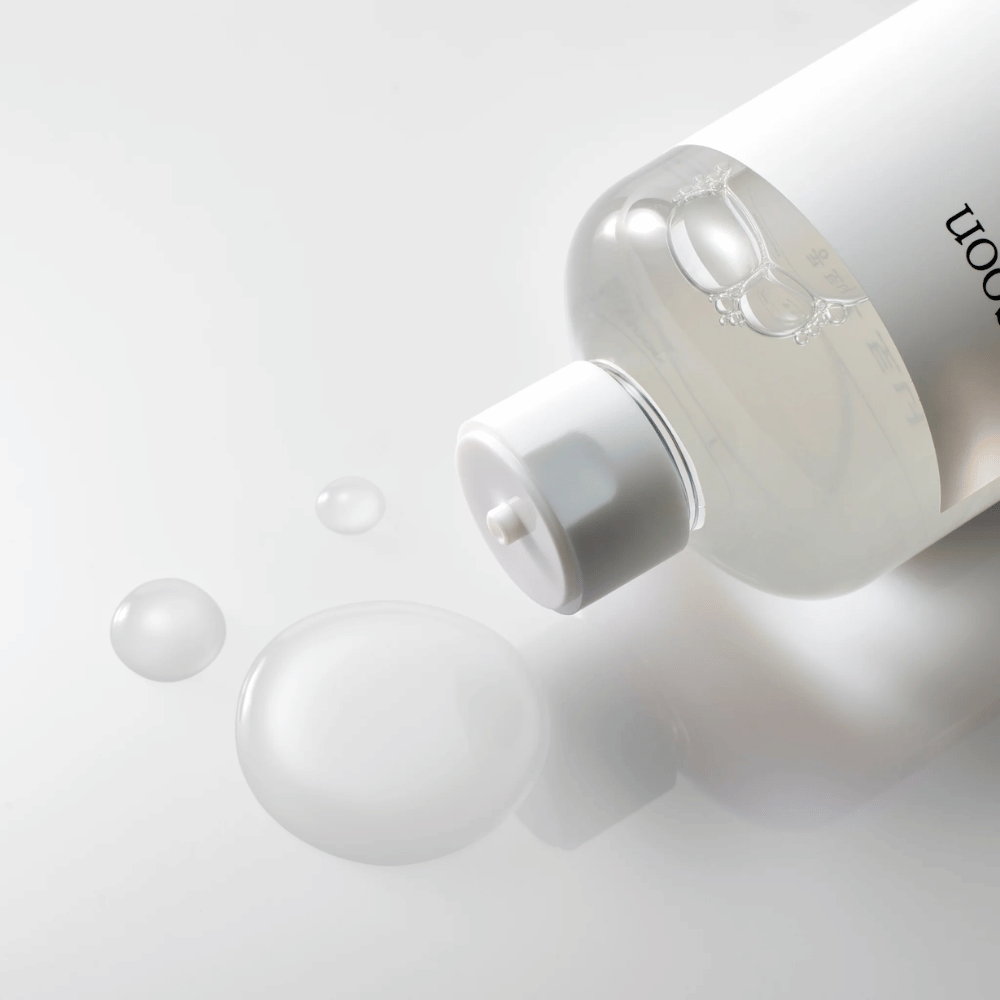 Une bouteille transparente est couchée sur le côté sur une surface brillante avec un toner léger qui s'écoule lentement et forme des gouttes et une plus grande flaque sur la surface. La bouteille a une étiquette blanche et un mécanisme de pompe blanc. L'éclairage est doux et crée une atmosphère propre et apaisante, tandis que la consistance fluide et la clarté du toner sont mises en valeur par la façon dont il se répand sur la surface.