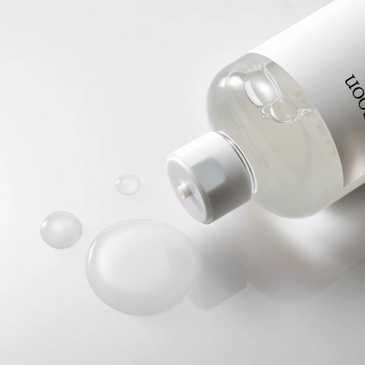 Une bouteille transparente est couchée sur le côté sur une surface brillante avec un toner léger qui s'écoule lentement et forme des gouttes et une plus grande flaque sur la surface. La bouteille a une étiquette blanche et un mécanisme de pompe blanc. L'éclairage est doux et crée une atmosphère propre et apaisante, tandis que la consistance fluide et la clarté du toner sont mises en valeur par la façon dont il se répand sur la surface.