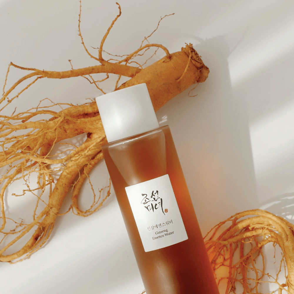 Une bouteille brune Beauty of Joseon Ginseng Essence Water avec un bouchon blanc est posée à côté d'une racine de ginseng fraîche sur fond blanc.