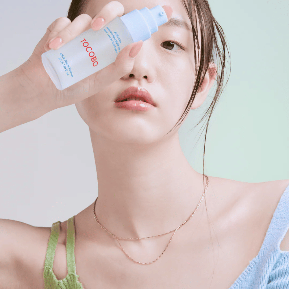 Jeune femme avec un maquillage doux et un teint naturel tenant une bouteille de TOCOBO Bifida Biome Essence devant son visage. Elle porte un pull bleu clair avec des bretelles vertes et un collier fin en or sur un fond clair.