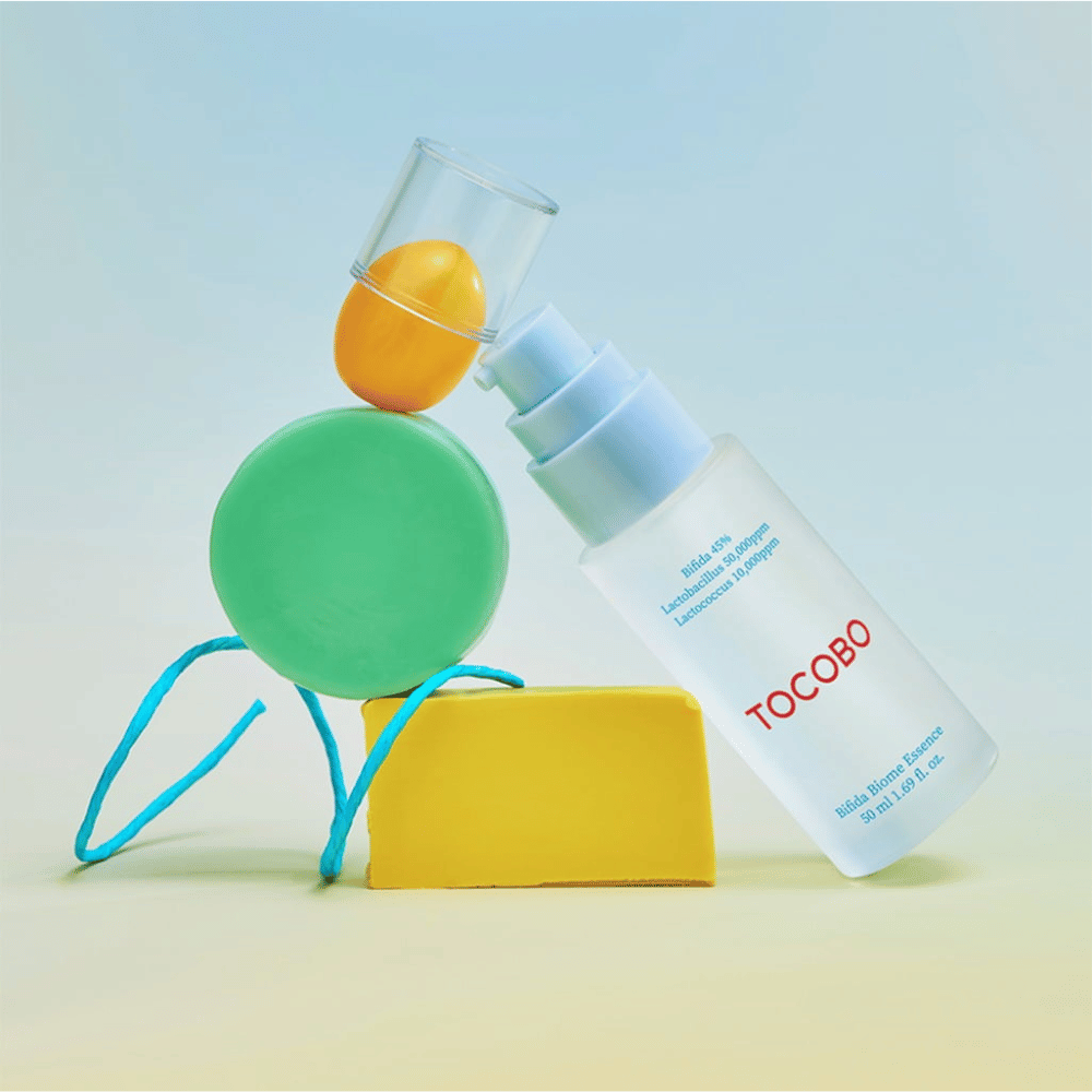 TOCOBO Bifida Biome Essence dans un flacon pompe givré avec un logo rouge, placé sur un fond coloré avec des formes géométriques vertes, jaunes et oranges. Le texte sur le flacon indique Bifida 5 %, Lactobacillus 3 000 ppm et Lactococcus 1 000 ppm.