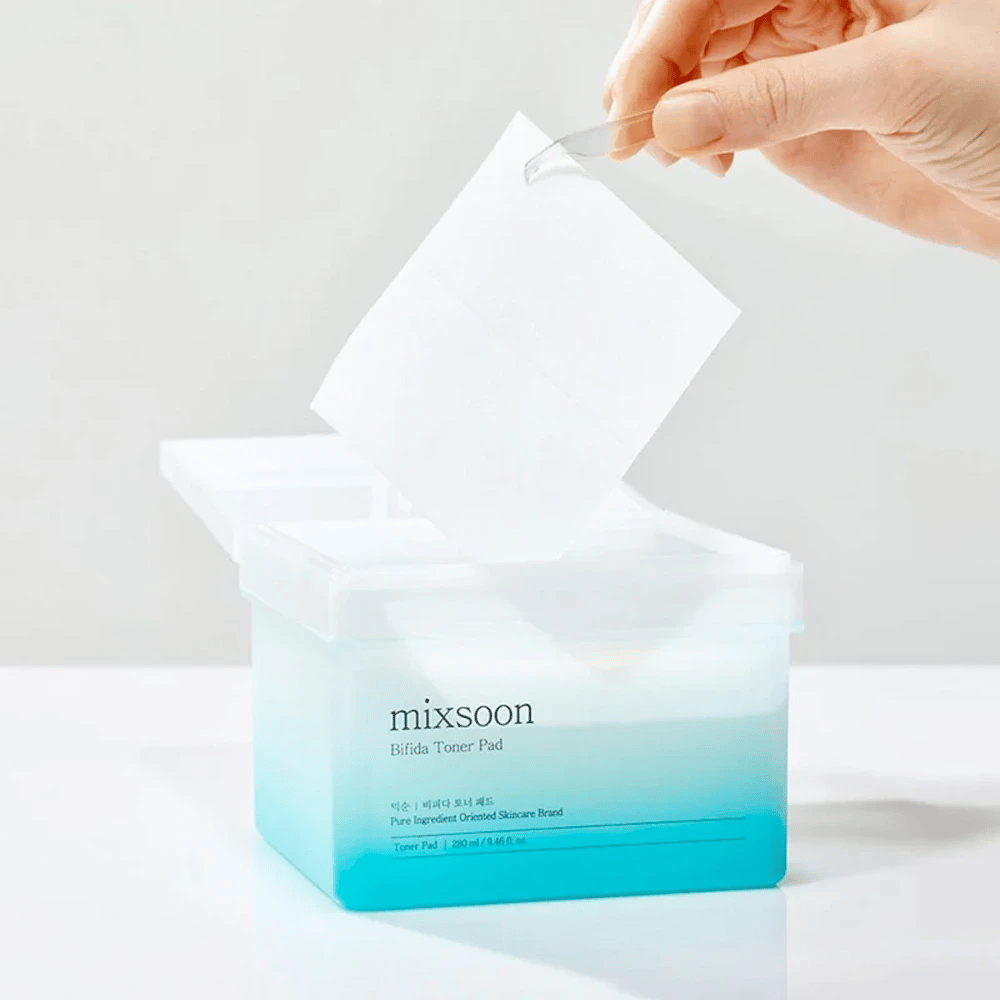 Un récipient transparent avec un dégradé bleu et blanc contenant des tampons de toner Bifida. Une main prend un tampon de toner du récipient, qui est étiqueté avec "mixsoon Bifida Toner Pad" et contient 280 ml (9,46 fl. oz.) de tampons de toner destinés au soin de la peau.