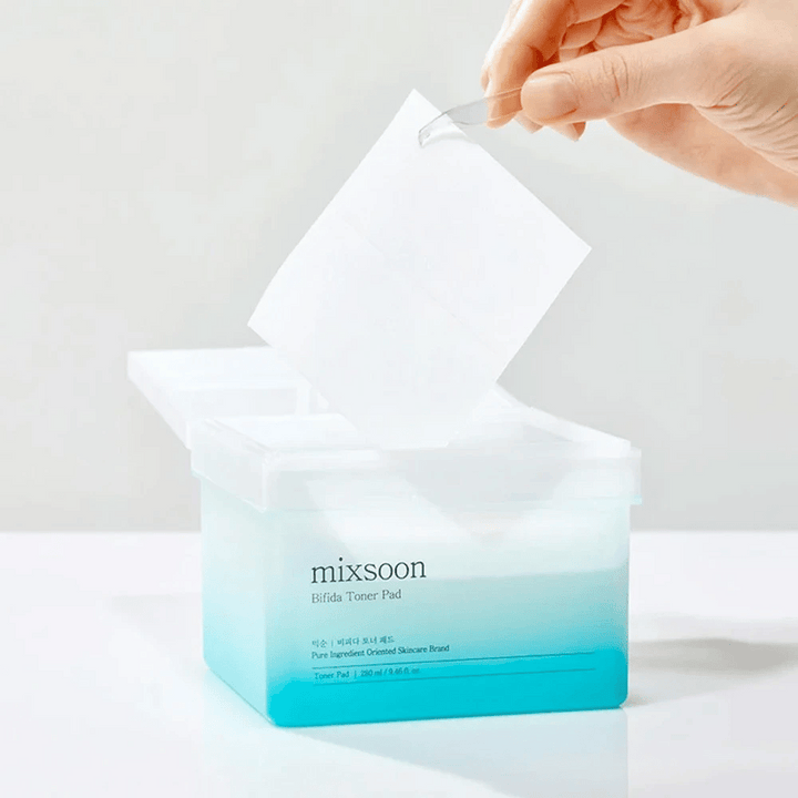 Un récipient transparent avec un dégradé bleu et blanc contenant des tampons de toner Bifida. Une main prend un tampon de toner du récipient, qui est étiqueté avec "mixsoon Bifida Toner Pad" et contient 280 ml (9,46 fl. oz.) de tampons de toner destinés au soin de la peau.