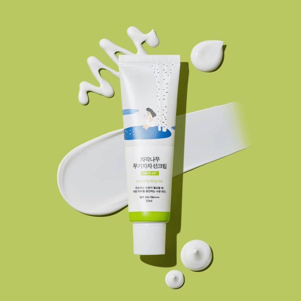 Birch Juice Moisturizing Mild Up Sunscreen – tube blanc avec base verte sur fond vert, entouré de crème solaire étalée et de gouttes, SPF 50+ PA++++, 50 ml.