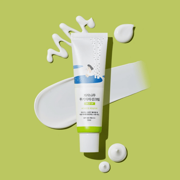 Birch Juice Moisturizing Mild Up Sunscreen – tube blanc avec base verte sur fond vert, entouré de crème solaire étalée et de gouttes, SPF 50+ PA++++, 50 ml.