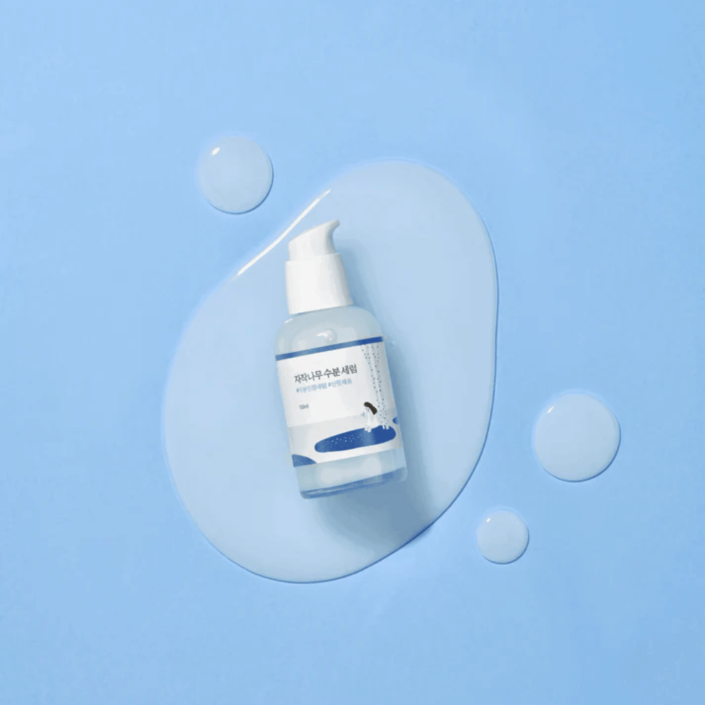 Round Lab Birch Juice Moisturizing Serum dans un flacon pompe transparent placé sur un fond bleu avec des gouttes et du sérum renversé.