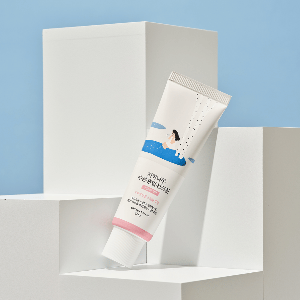 Un tube avec Birch Juice Moisturizing Tone Up Sunscreen placé sur des cubes blancs contre un fond bleu clair. Le tube est blanc avec une illustration d'une femme près d'un bouleau et de l'eau. Le texte en coréen indique que le produit a SPF 50+ PA++++ et contient 50 ml.