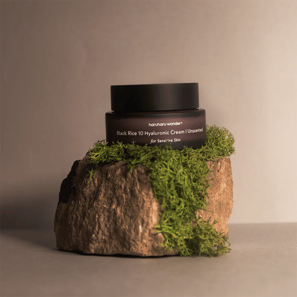 L'image montre un pot de crème pour la peau marqué "haruharu wonder Black Rice 10 Hyaluronic Cream Unscented for Sensitive Skin". Le pot est noir et repose sur une grande pierre couverte de mousse vert clair, créant une sensation naturelle et terre-à-terre.