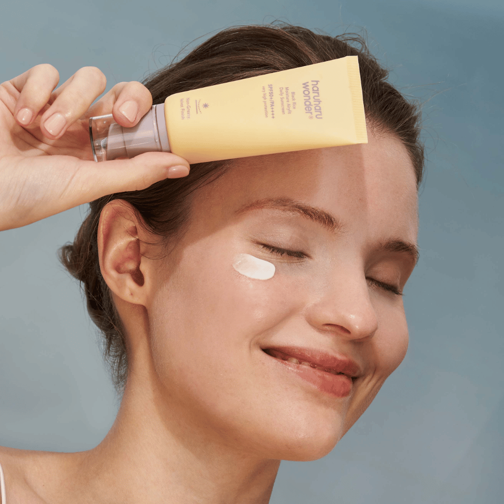 Une femme souriante applique Black Rice Moisture Airyfit Daily Sunscreen SPF 50+ PA++++ de Haruharu Wonder sur son visage. Elle tient un tube jaune de crème solaire et a une noisette du produit sur la joue. Le fond est bleu clair.