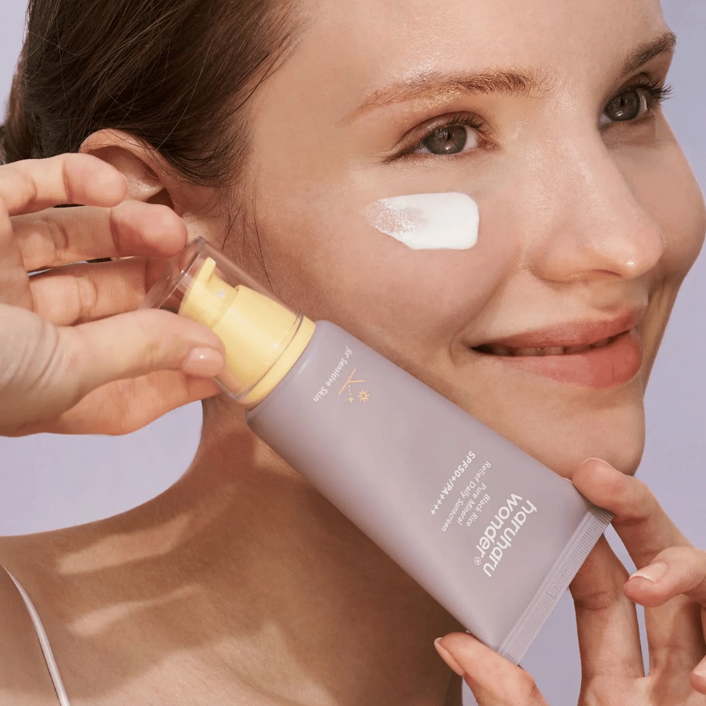Une femme souriante applique Haruharu Wonder Black Rice Pure Mineral Relief Daily Sunscreen SPF 50+ PA++++ sur son visage. Elle tient un tube violet de crème solaire et a une noisette du produit sur la joue. L'arrière-plan est lilas clair.