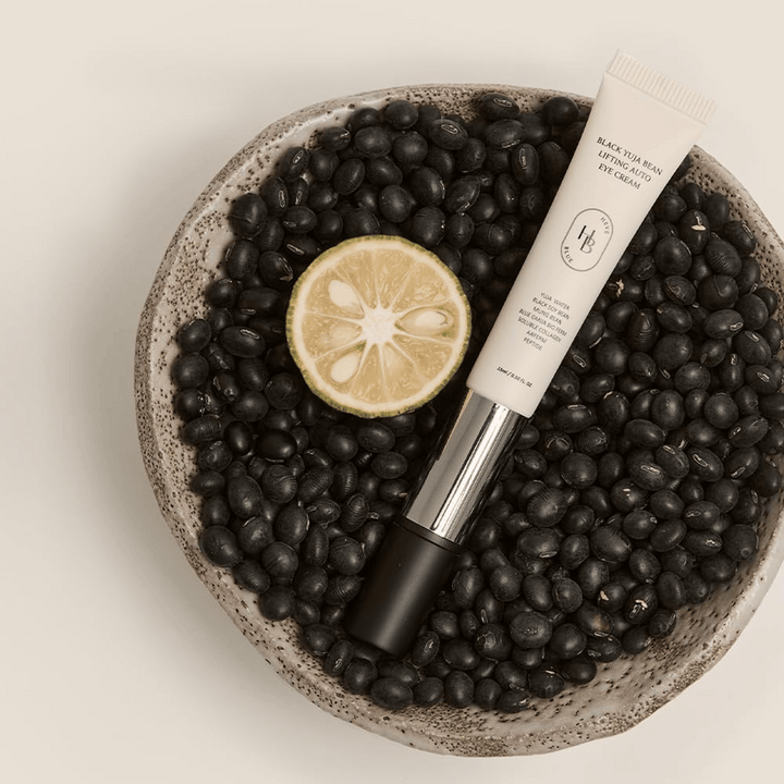 Black Yuja Bean Lifting Auto Eye Cream placé dans un bol rempli de haricots noirs et d'un agrume coupé en deux – une représentation visuelle des ingrédients naturels du produit.