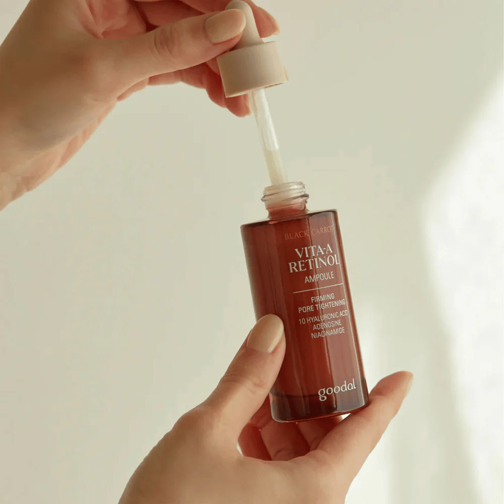 Une main tient une bouteille avec Goodal Black Carrot Vita-A Retinol Firming Ampoule. L'étiquette de la bouteille montre le nom et les propriétés du produit, y compris "Affermissement et Rétrécissement des Pores" et des ingrédients comme 10% d'acide hyaluronique, adénosine et niacinamide. La pipette est de couleur beige.