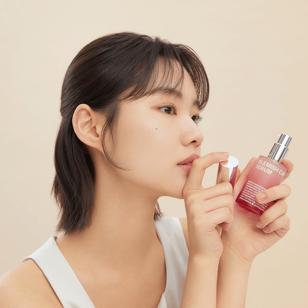 Une femme aux cheveux foncés tient une bouteille de Blemish Care Serum dans une main et enlève le bouchon avec l'autre. Elle a une expression neutre et le fond est beige clair, ce qui donne une impression douce et minimaliste.