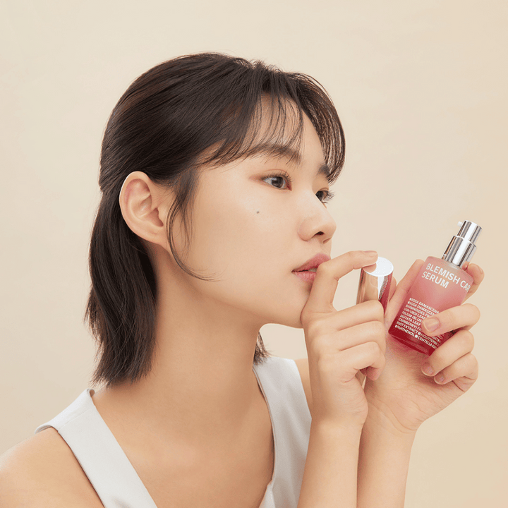 Une femme aux cheveux foncés tient une bouteille de Blemish Care Serum dans une main et enlève le bouchon avec l'autre. Elle a une expression neutre et le fond est beige clair, ce qui donne une impression douce et minimaliste.